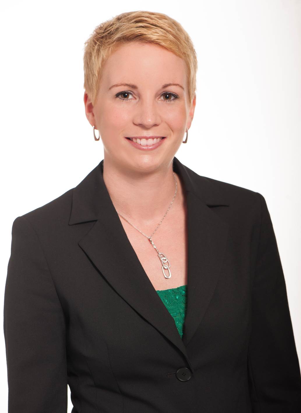 Christina Huber hat kurze blonde Haare, trägt Ohrringe, einen schwarzen Blazer mit grünem Shirt darunter.